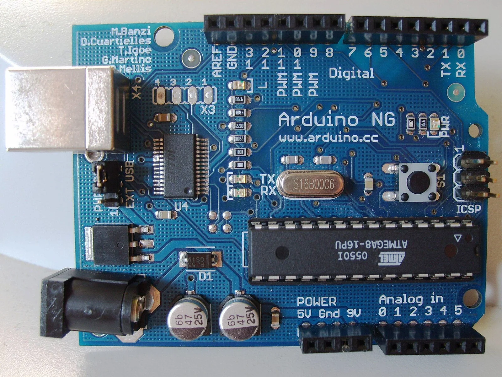 Vista dall'alto di una scheda Arduino dei primi anni con microcontrollore Atmel ATmega8, regolatore, quarzo, connettori per i pin di I/O e interfaccia seriale, su piano scuro.