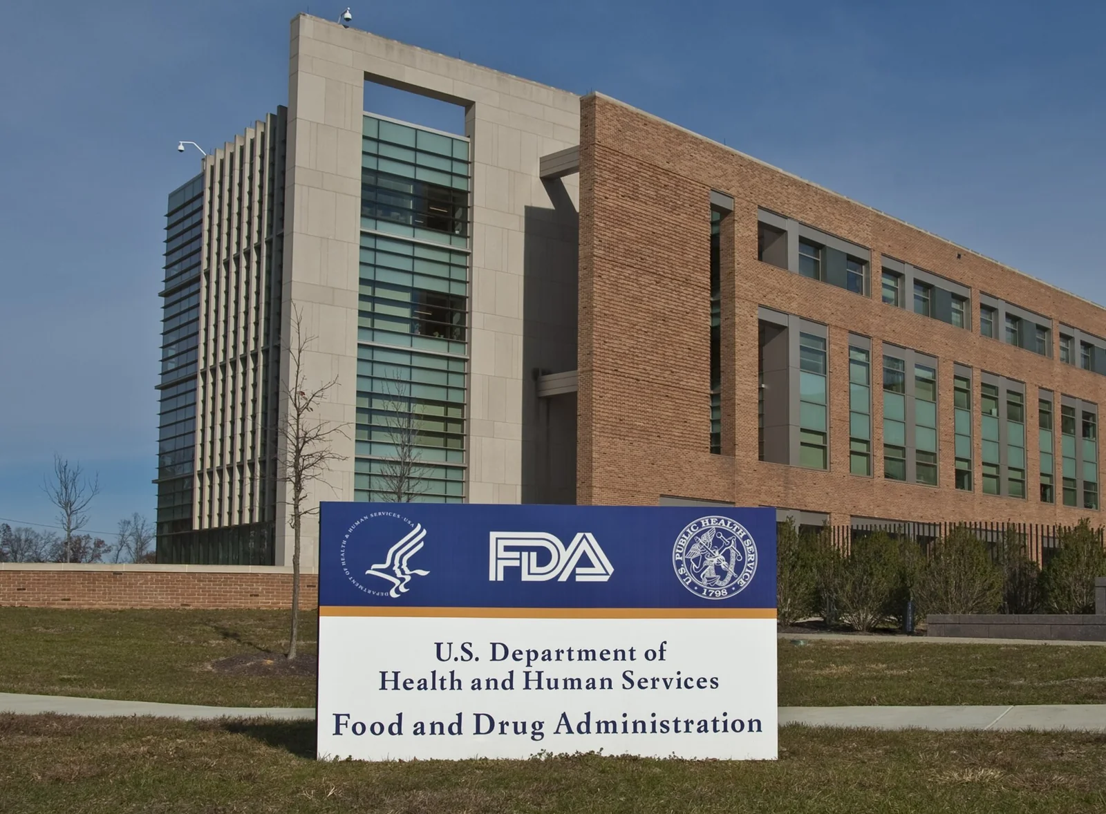 Cartello d'ingresso bianco con il logo della U.S. Food and Drug Administration al campus di White Oak; sullo sfondo l'edificio in mattoni e vetro della sede FDA.