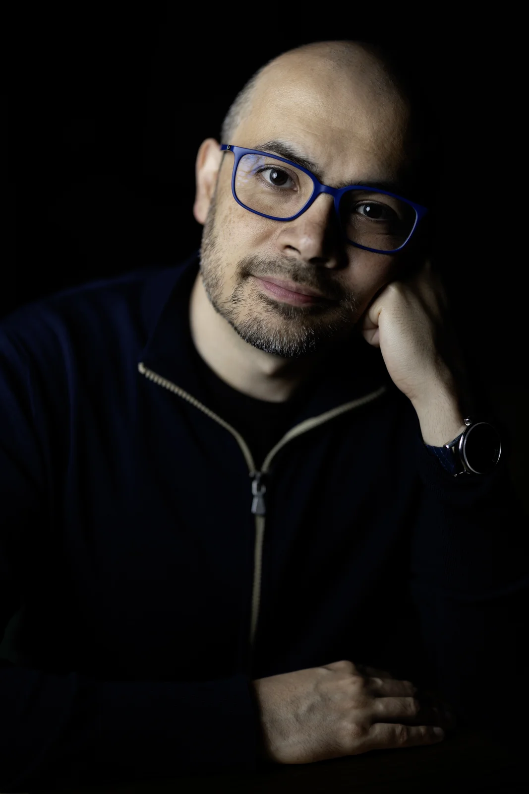 Ritratto di Demis Hassabis su fondo nero: occhiali dalla montatura blu, giacca scura con zip, il mento appoggiato alla mano destra (2025).