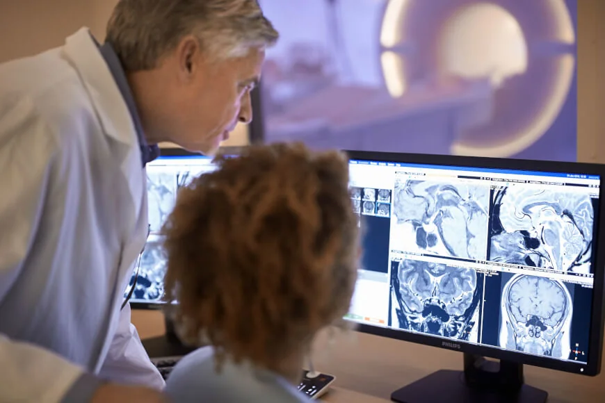 Un radiologo seduto a una postazione clinica interpreta immagini di risonanza magnetica del cervello visualizzate su un monitor medicale.