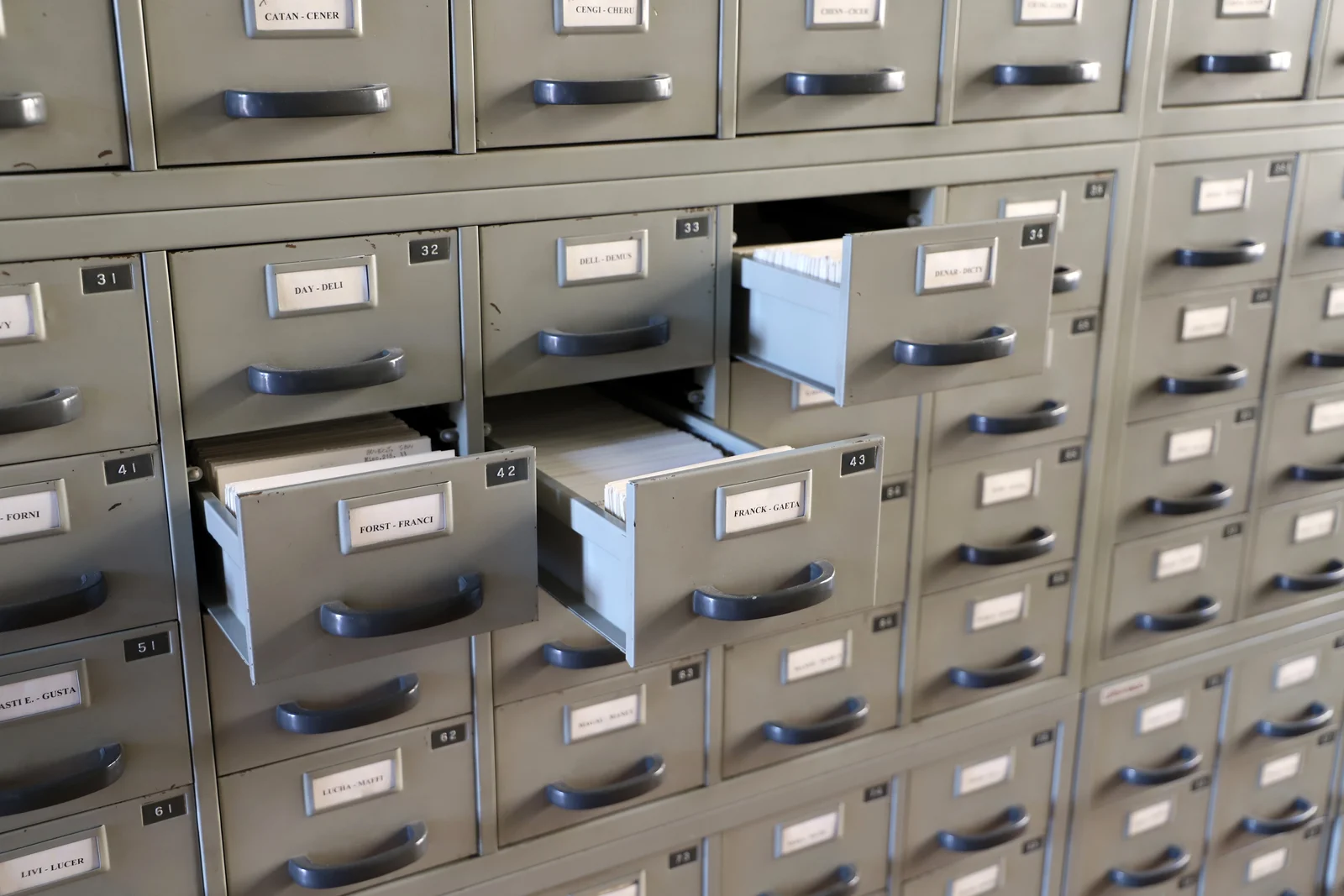 Schedario di una biblioteca: decine di cassetti metallici grigi con maniglie, ciascuno con un'etichetta che riporta l'intervallo alfabetico contenuto — CATAN-CENER, DAY-DELI, FORST-FRANCI. Tre cassetti sono aperti e lasciano vedere le schede di cartoncino allineate.