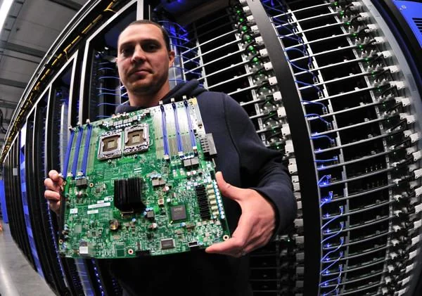 Un ingegnere di Facebook tiene in mano una scheda madre server in un data center, con file di rack sullo sfondo.