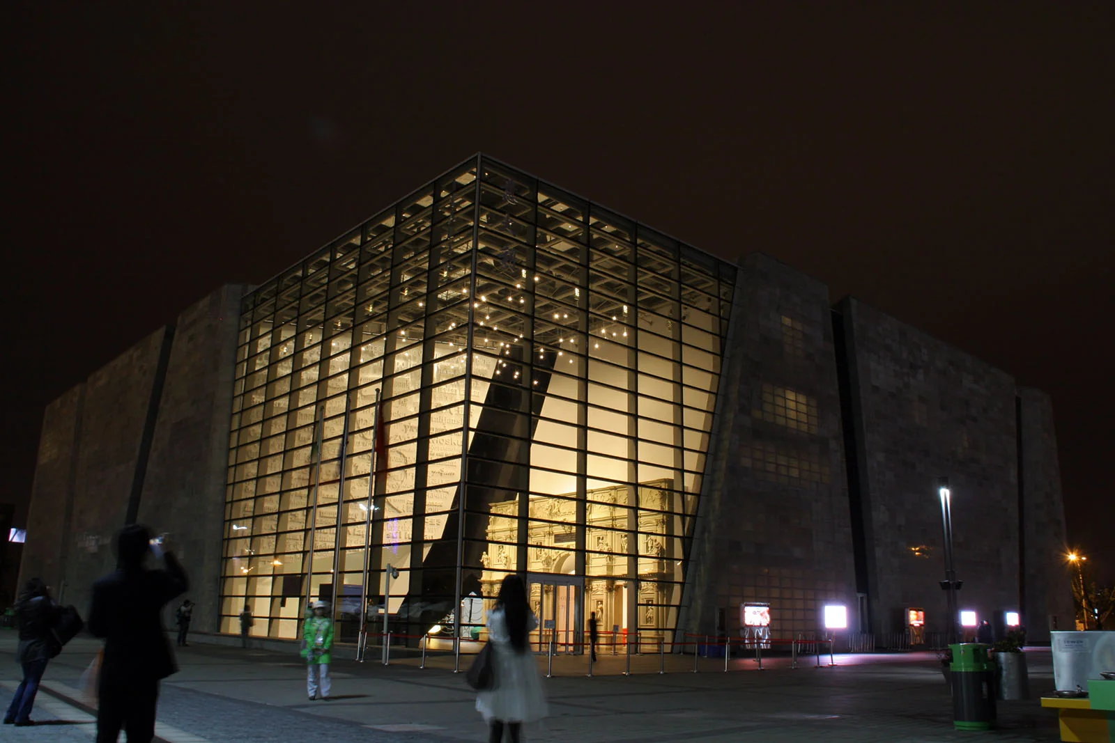 Il Padiglione Italia all'Expo 2010 di Shanghai di notte, con l'ampia facciata in vetro illuminata dall'interno e alcuni visitatori in primo piano.