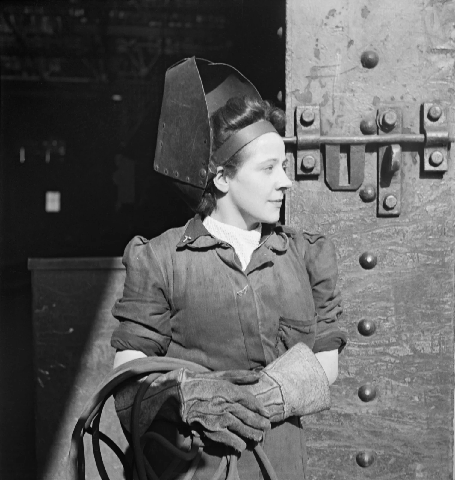 Fotografia in bianco e nero del 1943: una giovane saldatrice in un cantiere navale del Tyneside, con la maschera da saldatura sollevata sulla fronte, i guanti da lavoro e il cavo della torcia fra le mani, davanti a una parete di acciaio chiodato.